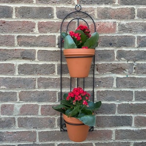 Peut inclure: Un porte-plantes mural en métal noir avec deux pots en terre cuite et des plantes à fleurs rouges.