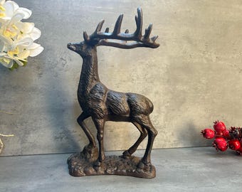 May include: A bronze-colored deer figurine with large antlers. The sculpture is standing on a small, textured base. The deer is posed in a lifelike stance, with detailed fur texture. The background includes white flowers and red berries.