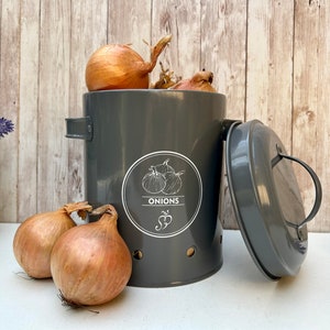 Peut inclure: Un récipient de rangement pour oignons en métal gris avec un couvercle et des trous d'aération. Le récipient porte une étiquette blanche sur laquelle est inscrit "ONIONS". Le récipient est rempli d'oignons bruns.