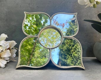 May include: A decorative silver mirror in a floral shape. The mirror has five sections, each reflecting trees and sky. The edges are decorated with small, beaded detailing. A stylish home decor item.