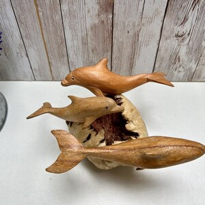 Peut inclure: Une sculpture en bois repr&eacute;sentant trois dauphins dans diff&eacute;rentes poses. Les dauphins sont sculpt&eacute;s dans du bois brun clair et semblent sauter d'une base textur&eacute;e de couleur claire. C'est un objet d&eacute;coratif.