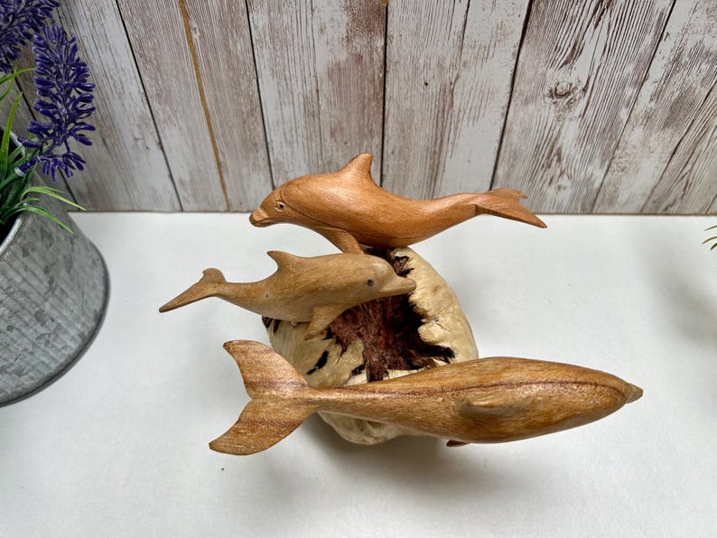 Peut inclure: Une sculpture en bois repr&eacute;sentant trois dauphins dans diff&eacute;rentes poses. Les dauphins sont sculpt&eacute;s dans du bois brun clair et semblent sauter d'une base textur&eacute;e de couleur claire. C'est un objet d&eacute;coratif.