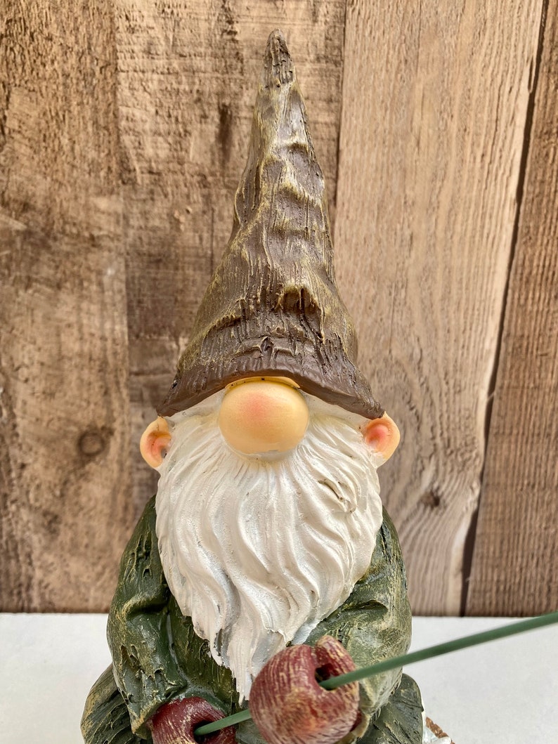 May include: A green and brown gnome figurine with a white beard and a pointed hat. The gnome is holding a fishing rod with a red bobber.