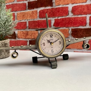 May include: A vintage-style aeroplane clock with a metal body and a round clock face. The clock face has black Roman numeral hour markers and gold hands. The clock is on a white surface.