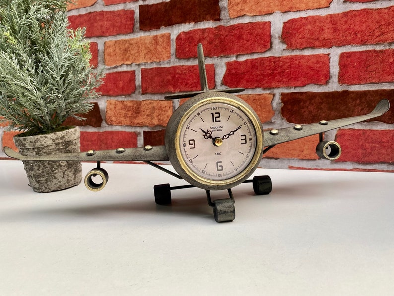 May include: A vintage-style aeroplane clock with a metal body and a round clock face. The clock face has black Roman numeral hour markers and gold hands. The clock is on a white surface.