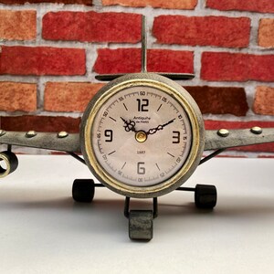 May include: A vintage-style aeroplane clock with a gold-tone finish and a white face. The clock features black Roman numeral hour markers and black hands. The clock is set on a white surface with a brick wall in the background.