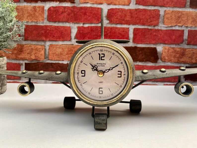 May include: A vintage-style aeroplane clock with a gold-tone finish and a white face. The clock features black Roman numeral hour markers and black hands. The clock is set on a white surface with a brick wall in the background.