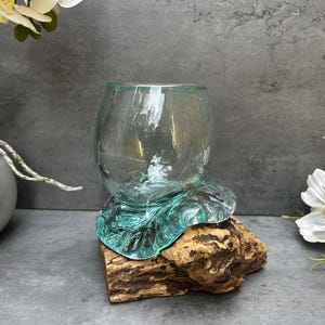 Peut inclure: Un bol en verre transparent est posé sur une base en verre de couleur turquoise, elle-même montée sur un morceau de bois naturel. Le bol est de forme arrondie. Le fond est un mur gris avec des fleurs blanches.