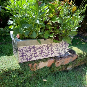 May include: A white planter with a purple floral pattern, filled with pink and orange flowers. The planter is sitting on a brick wall covered in moss.