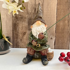 May include: A green and brown gnome figurine sitting on a rock with a fishing rod in hand. The gnome is wearing a green hat, a brown jacket, and brown boots. The gnome is holding a fishing rod with a green line. The gnome is sitting on a rock with a basket beside it.