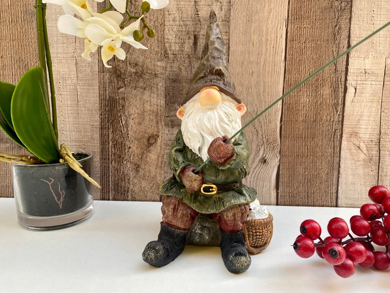 May include: A green and brown gnome figurine sitting on a rock with a fishing rod in hand. The gnome is wearing a green hat, a brown jacket, and brown boots. The gnome is holding a fishing rod with a green line. The gnome is sitting on a rock with a basket beside it.