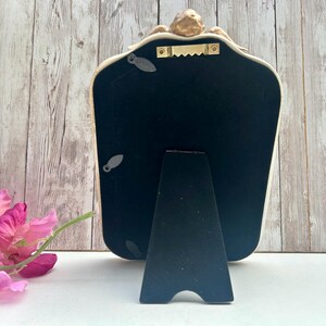 May include: A white and gold ornate mirror with a black stand. The mirror has a decorative design on the top and a black backing.
