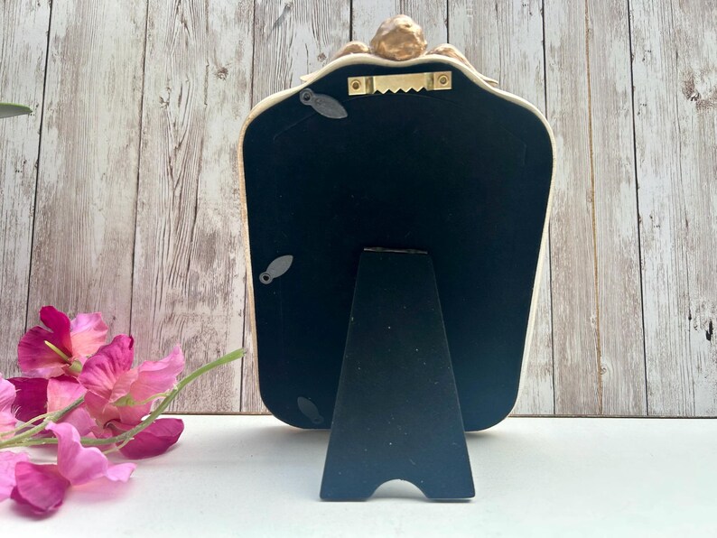 May include: A white and gold ornate mirror with a black stand. The mirror has a decorative design on the top and a black backing.