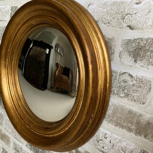 May include: A gold-toned round convex mirror with a wide, ornate frame. The mirror is reflecting a room with a fireplace and furniture.