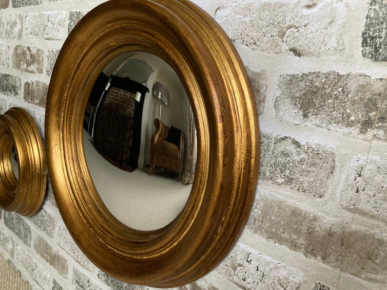 May include: A gold-toned round convex mirror with a wide, ornate frame. The mirror is reflecting a room with a fireplace and furniture.
