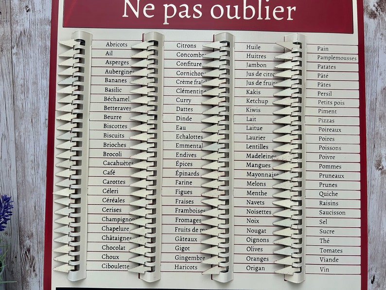 Op de afbeelding: Een witte boodschappenlijst met een rode kop die in het Frans "Ne pas oublier" zegt. De lijst is verdeeld in drie kolommen met verschillende voedingsmiddelen die in het Frans worden vermeld. De items zijn georganiseerd per categorie, zoals fruit, groenten en zuivelproducten.