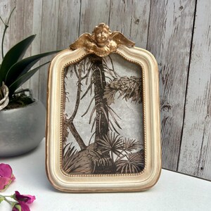 May include: A white picture frame with a gold cherub design on the top. The frame has a raised edge and a beaded border. The frame is empty and ready for a photo.