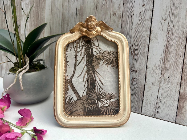 May include: A white picture frame with a gold cherub design on the top. The frame has a raised edge and a beaded border. The frame is empty and ready for a photo.