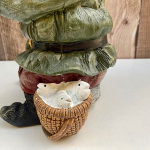 May include: A green and red gnome figurine holding a wicker basket with white fish inside. The gnome is wearing a green hat and a red and white belt.