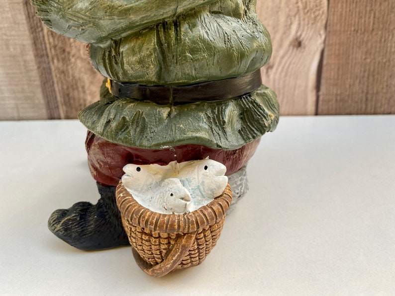 May include: A green and red gnome figurine holding a wicker basket with white fish inside. The gnome is wearing a green hat and a red and white belt.