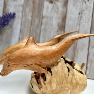 Peut inclure: Sculpture de dauphin en bois sculpt&eacute; &agrave; la main. Le dauphin est brun clair et repose sur une base en bois textur&eacute; brun clair. La sculpture est d&eacute;taill&eacute;e avec des nageoires et un museau pointu. Le fond est un panneau de bois clair.