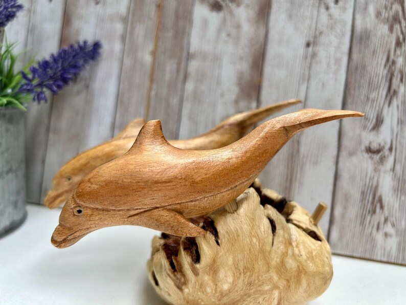Peut inclure: Sculpture de dauphin en bois sculpt&eacute; &agrave; la main. Le dauphin est brun clair et repose sur une base en bois textur&eacute; brun clair. La sculpture est d&eacute;taill&eacute;e avec des nageoires et un museau pointu. Le fond est un panneau de bois clair.