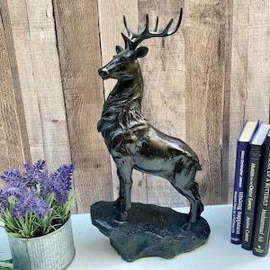 May include: A bronze-effect deer sculpture with detailed antlers and fur stands on a textured base. The statue is displayed near a small lavender plant in a metal container and several books with titles visible.