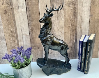 May include: A bronze-colored deer statue with detailed antlers and fur stands on a textured base. The sculpture is displayed near a small lavender plant in a metal container and several books with titles visible.