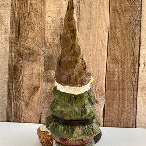 May include: A green and brown gnome figurine sitting on a rock with a fishing rod and a basket. The gnome is wearing a green hat and a green jacket with a brown belt.