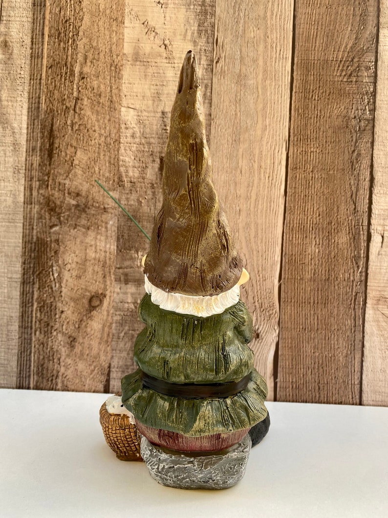 May include: A green and brown gnome figurine sitting on a rock with a fishing rod and a basket. The gnome is wearing a green hat and a green jacket with a brown belt.