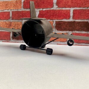May include: A grey metal aeroplane model with black wheels and a black propeller. The model is sitting on a white surface in front of a red brick wall.