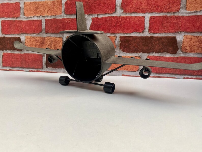 May include: A grey metal aeroplane model with black wheels and a black propeller. The model is sitting on a white surface in front of a red brick wall.