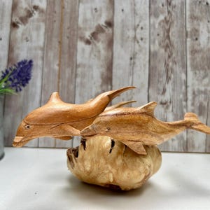 Peut inclure: Deux sculptures de dauphins en bois, avec une finition naturelle, sont pos&eacute;es sur une base textur&eacute;e de couleur claire. Les dauphins sont en plein saut, montrant leurs nageoires et leurs corps profil&eacute;s. L'arri&egrave;re-plan est un mur en planches de bois.
