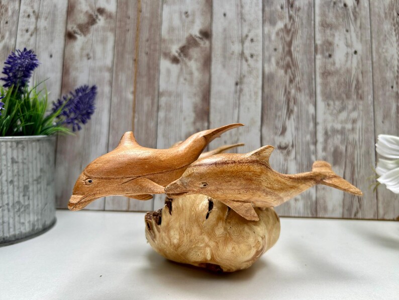 Peut inclure: Deux sculptures de dauphins en bois, avec une finition naturelle, sont pos&eacute;es sur une base textur&eacute;e de couleur claire. Les dauphins sont en plein saut, montrant leurs nageoires et leurs corps profil&eacute;s. L'arri&egrave;re-plan est un mur en planches de bois.