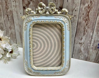 Peut inclure: Cadre photo rectangulaire orné, bleu clair avec des détails dorés. Il présente un nœud en haut et des bordures décoratives. L'intérieur affiche un motif circulaire beige et blanc, typique des cadres vintage.