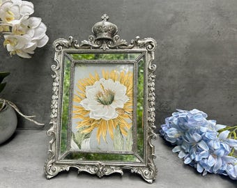 May include: Ornate silver-toned picture frame with a crown detail at the top. The frame holds a floral print with a white and yellow flower. Decorative elements include scrolling patterns. A beautiful home decor item.