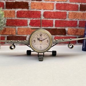 May include: A metal aeroplane clock with a round clock face and black hands. The clock is on a white surface with a brick wall in the background.