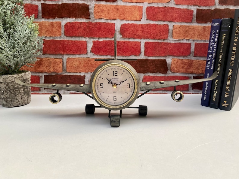 May include: A metal aeroplane clock with a round clock face and black hands. The clock is on a white surface with a brick wall in the background.