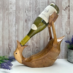 May include: A wooden wine bottle holder shaped like a bird, supporting a green wine bottle. The bird is crafted from light and dark brown wood. The wine bottle features a white label with black text. The backdrop is a light wood panel.