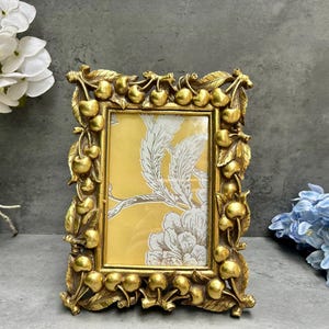 Peut inclure: Un cadre photo rectangulaire doré avec un motif de cerises et de feuilles. Le cadre contient une impression florale jaune et blanche. Cadre décoratif pour afficher des photos ou des œuvres d'art.
