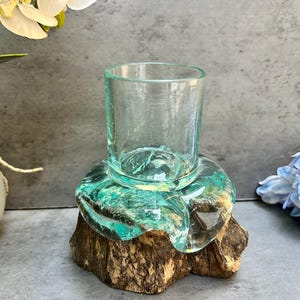 Peut inclure: Un gobelet en verre transparent posé sur un socle décoratif en résine turquoise et en bois naturel. Le socle a une forme organique unique. Le gobelet est vide et semble être fait de verre recyclé. L'esthétique générale est rustique et naturelle.