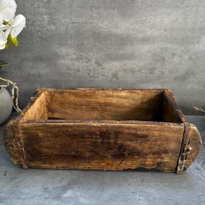 May include: A rectangular, weathered wooden brick mould with a dark brown, distressed finish. The box features visible wood grain, a rough texture, and a thin, rusted metal band wrapped around the exterior sides, showing signs of age and wear.