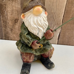 May include: A green and brown gnome figurine with a white beard and a red hat is sitting on a rock and holding a fishing rod.