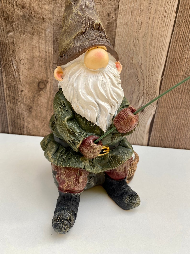 May include: A green and brown gnome figurine with a white beard and a red hat is sitting on a rock and holding a fishing rod.