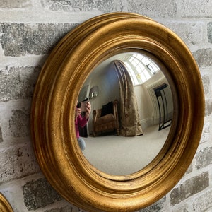 May include: A gold-framed round mirror with a convex lens. The mirror is hanging on a white brick wall.