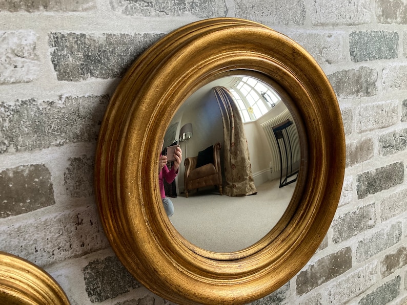 May include: A gold-framed round mirror with a convex lens. The mirror is hanging on a white brick wall.