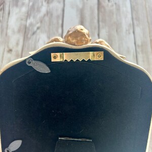 May include: The back of a gold-colored picture frame with a black velvet backing. The frame has a decorative design and a hanging hook.