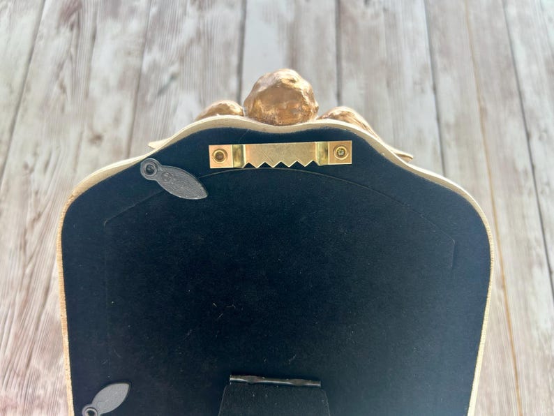 May include: The back of a gold-colored picture frame with a black velvet backing. The frame has a decorative design and a hanging hook.