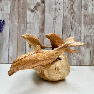 Peut inclure: Une sculpture en bois repr&eacute;sentant trois dauphins sautant d'une base de couleur claire. Les dauphins sont sculpt&eacute;s dans un bois brun clair, avec un grain et des d&eacute;tails visibles. La sculpture est pos&eacute;e sur un fond en bois.