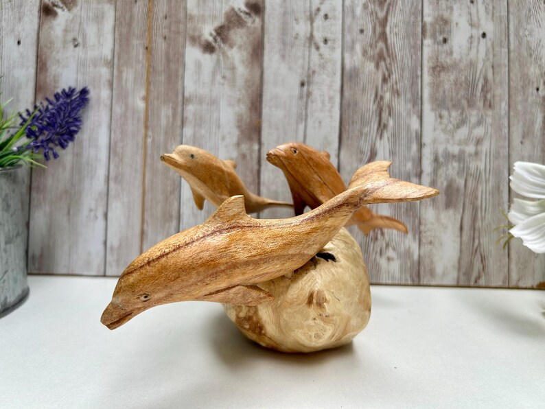 Peut inclure: Une sculpture en bois repr&eacute;sentant trois dauphins sautant d'une base de couleur claire. Les dauphins sont sculpt&eacute;s dans un bois brun clair, avec un grain et des d&eacute;tails visibles. La sculpture est pos&eacute;e sur un fond en bois.